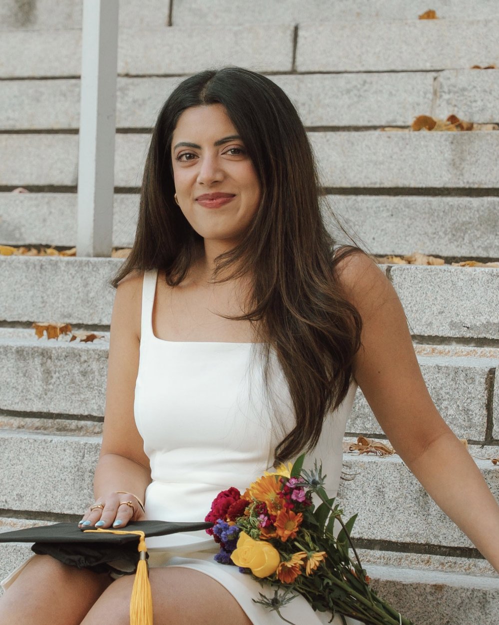 Yuvika smiling on stone steps, holding a graduation cap and flowers